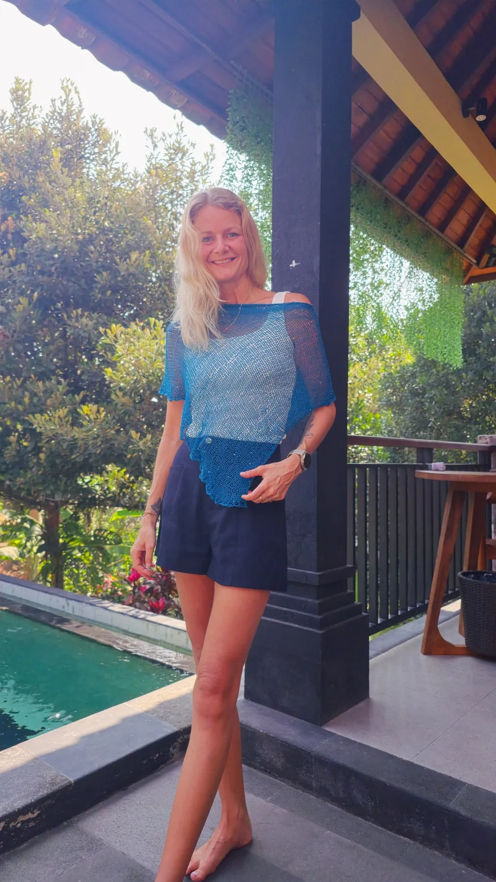 Tine står barbeint og smiler i Embun Poncho, lent mot en mørk søyle ved bassenget. Det blå, luftige strikket faller ned i en spiss foran, over mørke shorts.