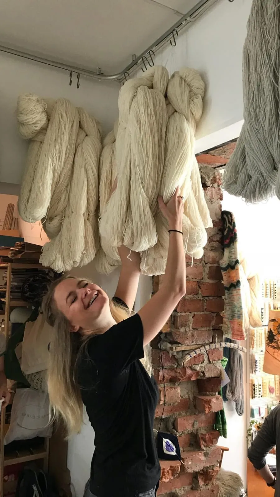 Tine strekker armene opp mot et tak fullt av store ullgarn-nøster i en garnbutikk, og ler.