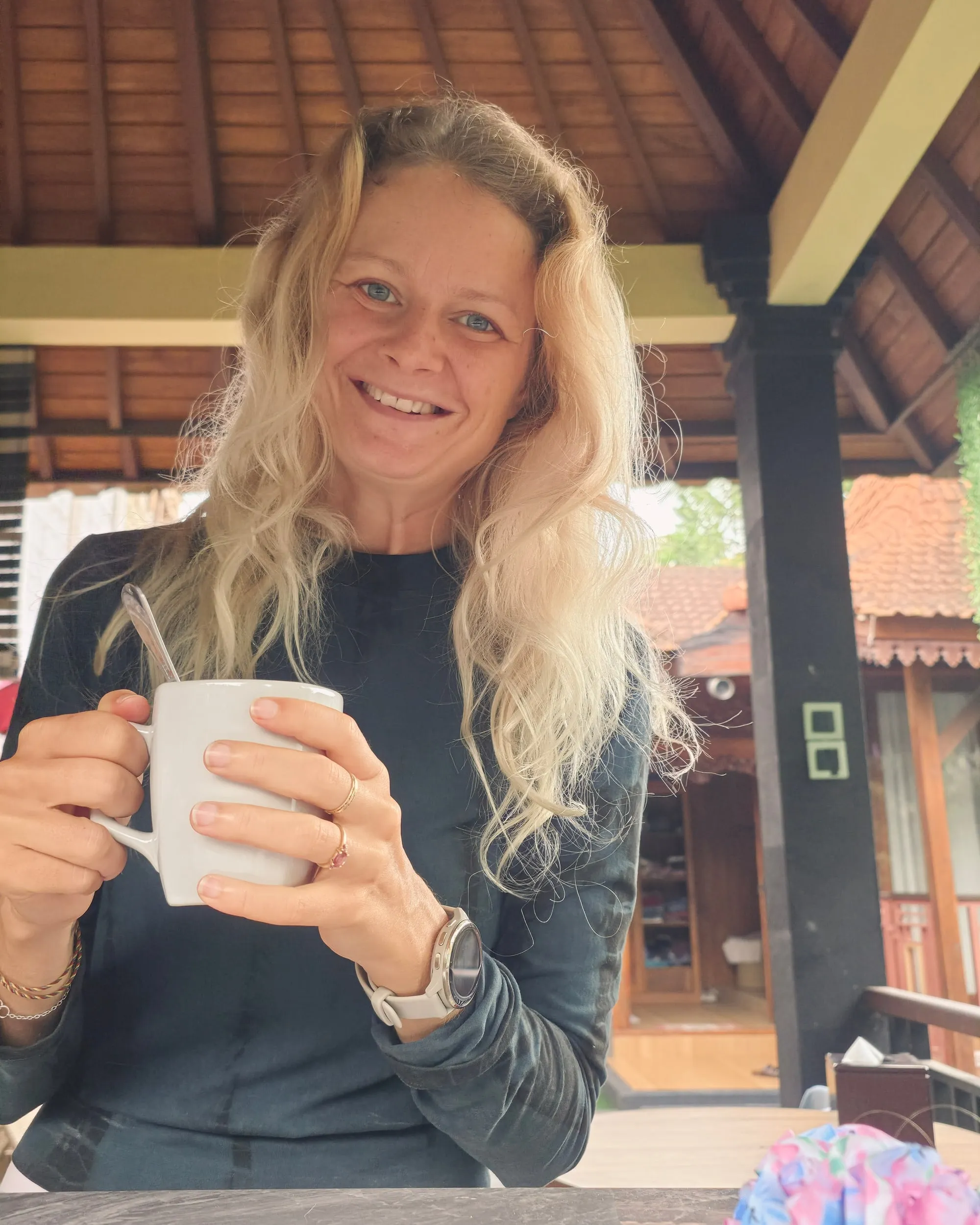 Tine holder koppen med begge hender og smiler, under tretaket med grønt og tegltak bak.