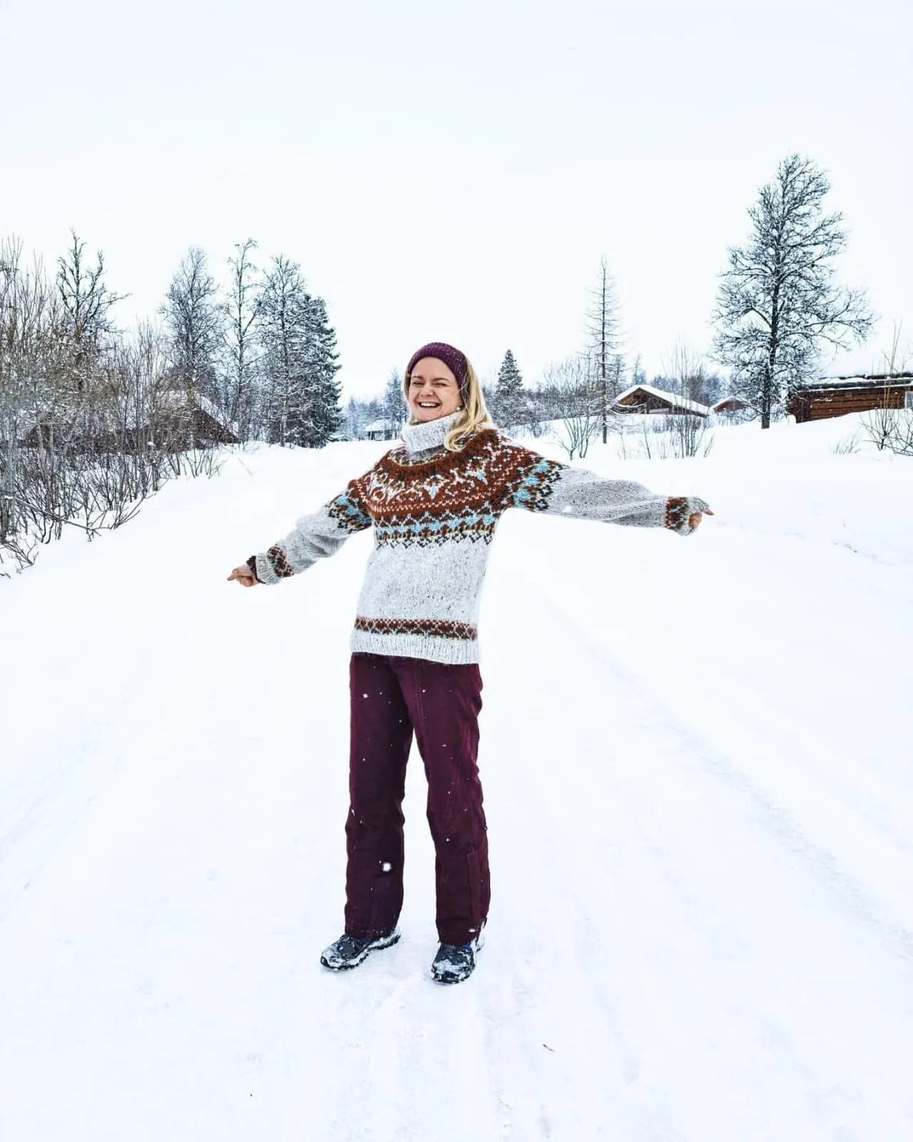 Tine står midt på en snødekt vei i Norge med armene ut til siden, i mønsterstrikket ullgenser.