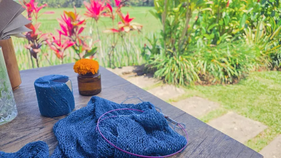 Ponchoen på rundpinne i blått garn, på et bord med oransje blomster ved siden av.
